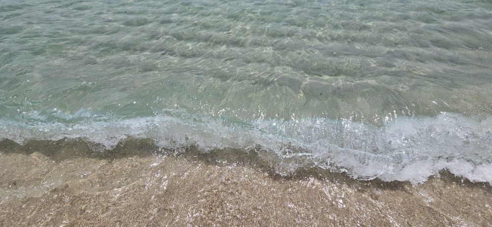 The image shows a serene shoreline where gentle waves of crystal-clear water meet the sandy beach.