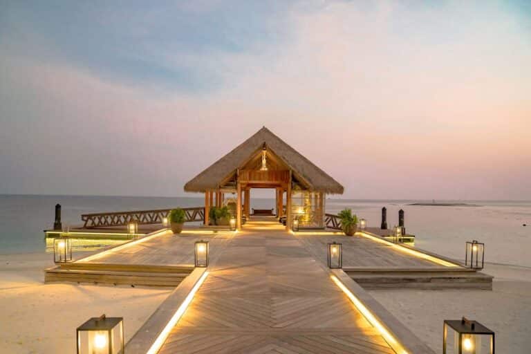 Luxurious beachfront pavilion in the Maldives at sunset, surrounded by tranquil ocean waters and a lit pathway extending toward the sea.